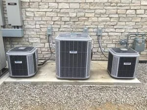 Three black AC condenser units installed on gravel pad against brick wall