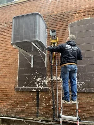 Technician installing large commercial HVAC unit on brick building with ductwork and electrical connections