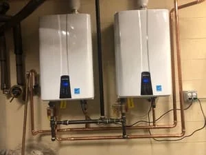 Two white tankless water heaters with digital displays mounted on wall with copper piping and brass manifold connections